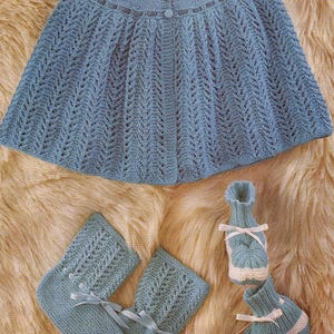 Peut inclure: Ensemble de bébé tricoté bleu clair, comprenant une cape boutonnée, des chaussettes avec des rubans et des chaussons avec une bordure blanche. L'ensemble est présenté sur une surface texturée brun clair.