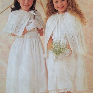 May include: Two young girls wearing white dresses and lace capes. The girl on the left is holding a white parasol and the girl on the right is holding a bouquet of white flowers.
