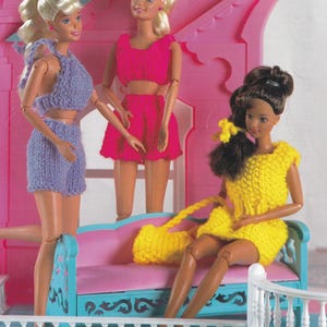 May include: Three fashion dolls are posed in a pink and blue dollhouse setting. The dolls are wearing knitted outfits in purple, pink, and yellow. One doll is seated on a pink sofa with a yellow handbag.
