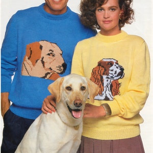 May include: A couple wearing matching sweaters with dog illustrations. The man is wearing a blue sweater with a brown and tan Labrador Retriever. The woman is wearing a yellow sweater with a brown and white spaniel. A white Labrador Retriever is sitting in front of them.