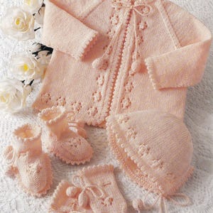 May include: A pale pink knitted baby set, including a jacket, booties, mittens, and a bonnet. The set features floral detailing and pom-pom accents. The jacket has a tie closure.