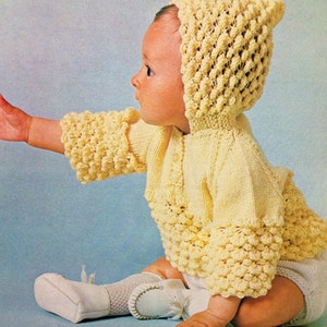 May include: A baby wearing a yellow knitted hooded sweater and matching booties. The sweater and booties have a textured, bobbly design.