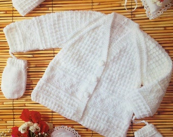 May include: A white knitted baby set, including a cardigan, hat, and mittens. The set is made of a textured knit fabric and is displayed on a bamboo mat. Decorative pillows and flowers are also present.
