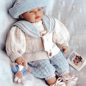 Puede incluir: Muñeca vestida con un conjunto de punto en azul claro y crema. El conjunto incluye un gorro, un jersey de estilo marinero y un pantalón. La muñeca lleva botines de punto con lazos rosas y sostiene un pequeño peluche.