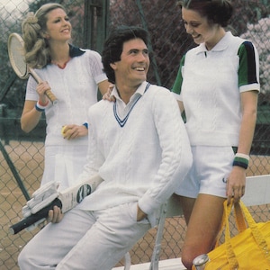 May include: Three people wearing white tennis outfits are standing on a tennis court. The man is wearing a white sweater with a blue stripe around the neck. The woman on the left is holding a tennis racket. The woman on the right is holding a yellow bag.