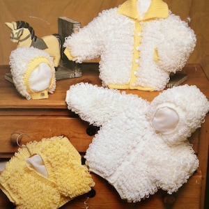 Könnte beinhalten: Vier gestrickte Babyjacken in Weiß und Gelb. Die Jacken sind aus einem flauschigen, strukturierten Garn gefertigt. Zwei der Jacken haben Kapuzen und eine hat einen Kragen.