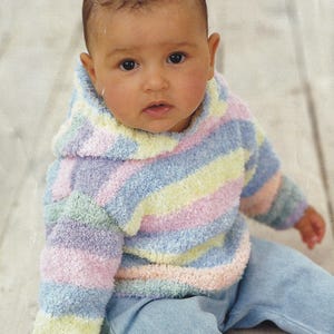 May include: A baby wearing a pastel striped hooded sweater and light blue jeans. The sweater features horizontal stripes in shades of pink, yellow, blue, and green. The baby is sitting on a light-colored wooden surface.