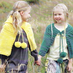 May include: Two young girls in a field, holding hands and wearing knitted capes. One wears a yellow hooded cape with pom-poms, the other a green cape with a tie and pom-poms. The capes appear handmade.