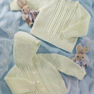 Può includere: Due maglioni per neonati in maglia giallo chiaro e un cardigan con cappuccio e bottoni perlati sono esposti su una superficie blu. Due coniglietti di peluche con fiocchi blu sono posizionati vicino ai maglioni. I maglioni hanno motivi strutturati.