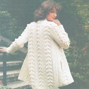 Könnte beinhalten: Cremefarbener, langer Strickmantel mit strukturiertem Muster. Der Mantel hat eine Reihe vertikaler strukturierter Säulen und Noppendetails. Die Ärmel sind lang und der Mantel hat eine ausgestellte Form. Die Frau steht auf einer Außentreppe.