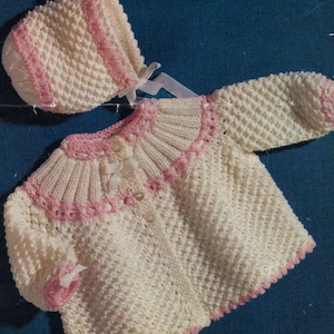 May include: A knitted baby cardigan and hat set in white and pink. The cardigan has a scalloped collar and button closure. The hat has a pink trim.
