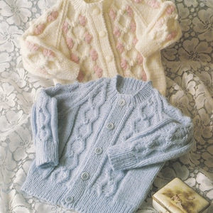 May include: Two knitted baby cardigans. One is light blue with a cable knit design and button closure. The other is cream-colored with pink floral accents and button closure. Both are on a white lace surface.