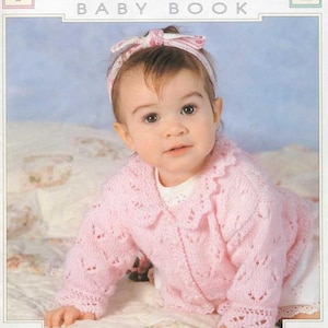 May include: A pink knitted baby cardigan with a lace pattern and a white collar. The cardigan is worn by a baby with a pink bow in their hair. The baby is crawling on a white and pink blanket. The text "TRADITIONAL BABY BOOK" is at the top of the image.