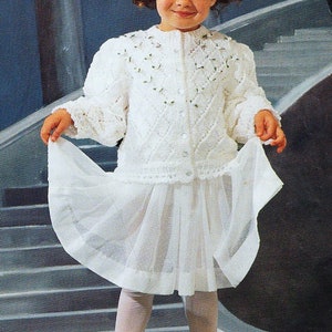 May include: A white knitted cardigan with a delicate floral pattern and a white skirt. The cardigan has a button closure and long sleeves. The skirt is made of a sheer fabric and has a ruffled hem.