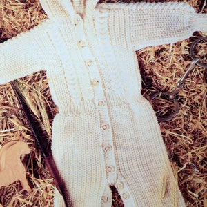 May include: Cream-colored, hooded baby jumpsuit with button closures. The knitted garment features a ribbed texture and long sleeves. The jumpsuit is displayed on a bed of straw, with a wooden toy, a whip, and a metal horse bit.