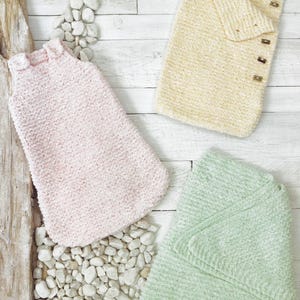 May include: Three knitted items: a sleeveless pale pink garment with shoulder straps, a rectangular pale yellow wrap with four brown buttons along the side, and a folded pale green textured blanket. All items feature a soft, nubby yarn texture.