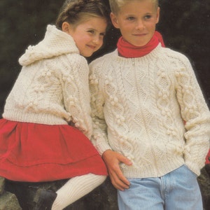 May include: Two children wearing cream-colored knitted sweaters with a cable knit pattern and pom-poms. The girl is wearing a red skirt and the boy is wearing blue jeans.
