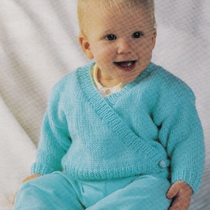 Könnte beinhalten: Ein Baby in einem hellblauen Strick-Wickelpullover und passender Hose. Der Pullover hat lange Ärmel und einen Knopfverschluss. Die Hose hat gelbe Bündchen an den Knöcheln. Das Baby lächelt.