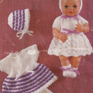 Puede incluir: Conjunto de ropa de punto para muñeca en blanco y morado. El conjunto incluye un vestido, un gorro y botines. El vestido tiene un corpiño blanco y una falda a rayas moradas y blancas. El gorro es blanco con rayas moradas. Los botines son blancos con lazos morados.