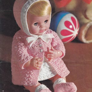 May include: A vintage doll wearing a pink knitted outfit, including a hooded jacket with white trim, a white dress, and matching booties. The doll has blonde hair and blue eyes. A white bow adorns the jacket. A colorful ball is in the background.