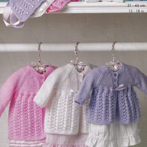 May include: Three knitted baby cardigans in pink, white, and lavender, hanging on decorative hangers. The cardigans have long sleeves and button closures. The measurements are 12-18 inches.