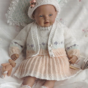 May include: A doll dressed in a knitted ensemble: a white cardigan with pastel details, a skirt, and a hat adorned with a pink flower. The doll also has matching shoes and a small teddy bear.