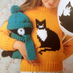 May include: A yellow knitted sweater with a black cat design and a white chest. The sweater is worn by a child holding a teal knitted hat and scarf.