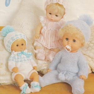 May include: Two knitted dolls wearing handmade outfits. The doll on the left is wearing a white and blue knitted outfit with a blue pom-pom hat. The doll on the right is wearing a light blue knitted outfit with a blue pom-pom hat. Both dolls have blonde hair and are sitting on a white blanket.