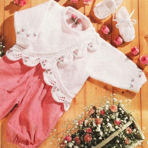 May include: A white knitted baby cardigan with a scalloped edge and floral embroidery. The cardigan is laid out on a wooden surface with a pair of white baby shoes, a basket of white flowers, and pink roses.