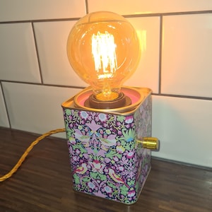 May include: A decorative table lamp featuring a large, round, amber light bulb. The square tin lamp base has a floral and bird pattern in pink, green, and blue. A gold-colored knob is on the side. The lamp is approximately 15 cm tall.