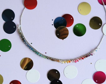 May include: A silver necklace with a rainbow of small beads. The necklace is surrounded by colorful sequins in various shades of red, green, gold, and white.
