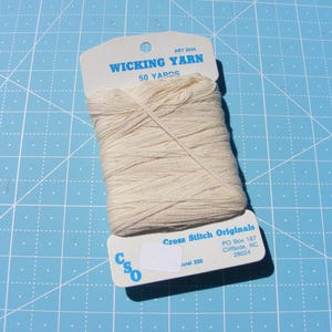 May include: A skein of off-white wicking yarn wrapped around a white cardboard card. The card features blue text reading WICKING YARN, 50 YARDS, ART 2034, Cross Stitch Originals, and CSO, along with a blank white sticker and address information.
