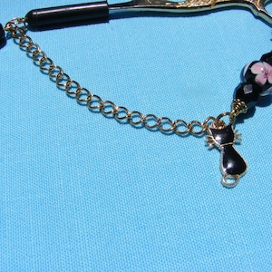May include: A gold-toned chain with a black cat charm, black and pink beads, and a black handle. The chain is attached to a pair of gold-toned scissors. The background is a light blue color.