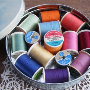 May include: A collection of colorful spools of thread inside a round metal tin. The spools are various colors, including orange, blue, pink, and green. The tin is partially open, revealing the thread inside. The tin is sitting on a white lace doily.