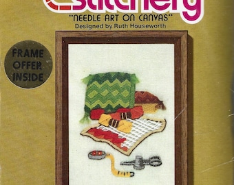 May include: A Jiffy Stitchery needlepoint kit featuring a framed design with a green pillow, a red and yellow blanket, and other needlepoint supplies. The kit includes the design, yarns, needle, and instructions. The package is labeled "Needle Art on Canvas."