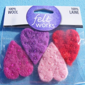 May include: A package of four felt hearts, each with a different color: purple, pink, red, and light pink. The hearts are decorated with small, shiny beads. The package is labeled "felt works" and "100% wool".