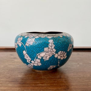 May include: A small, round, turquoise-colored decorative bowl with a floral design. The bowl features white and pink flowers and leaves, with a black rim and base. The exterior has a geometric pattern.
