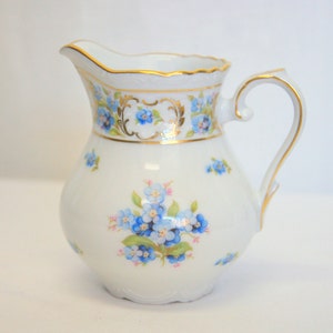 May include: White porcelain pitcher with a rounded body and a curved handle. The pitcher is decorated with blue floral patterns and gold trim around the rim and handle. A cluster of blue flowers is painted on the front.