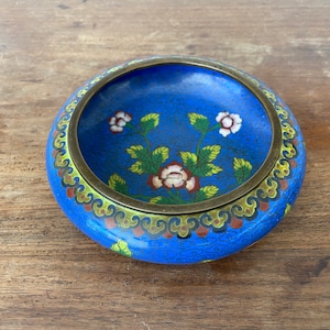 May include: A round, blue cloisonné dish with a floral design. The dish features a brass rim and a vibrant blue enamel background. The flowers are white and red with green leaves. A decorative piece.