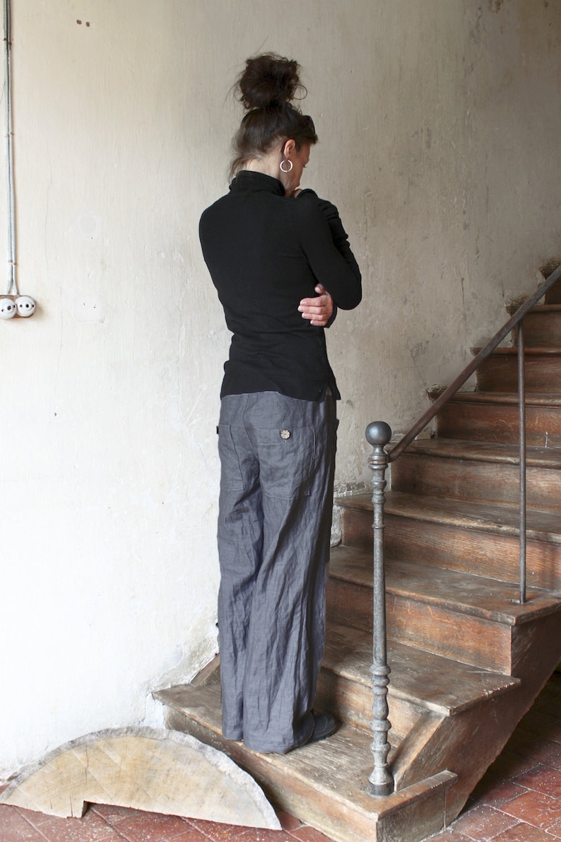 Puede incluir: Una mujer con un su&eacute;ter negro de cuello alto y pantalones de lino grises se encuentra de pie en una escalera de madera con una barandilla de metal.