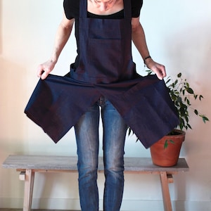 Puede incluir: Un delantal azul marino oscuro con escote cuadrado, tirantes anchos y un bolsillo frontal grande. El delantal presenta un diseño con abertura frontal que se ensancha en la cintura. El texto 'bloem linen' está impreso en negro en la esquina superior izquierda.