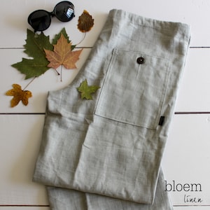 Puede incluir: Un pantal&oacute;n de lino gris claro con un gran bolsillo de parche en el lado derecho. El bolsillo tiene un cierre de bot&oacute;n. El pantal&oacute;n est&aacute; doblado y colocado plano sobre una superficie de madera blanca. Hay varias hojas de oto&ntilde;o y un par de gafas de sol en la superficie. El texto "bloem linen" es visible en la esquina inferior derecha de la imagen.