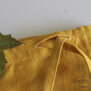Puede incluir: Primer plano de una bolsa de cord&oacute;n de lino amarillo con un cord&oacute;n anudado. La bolsa est&aacute; hecha de una tela suave y texturizada y tiene un dise&ntilde;o simple y elegante. El texto "bloem linen" es visible en la esquina inferior derecha de la imagen.