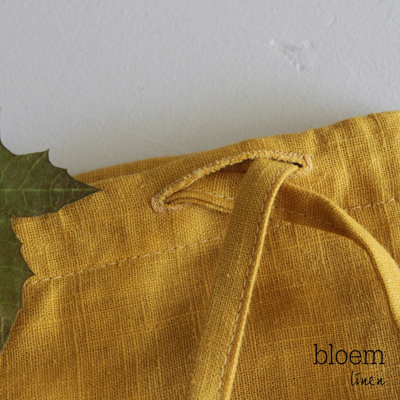 Puede incluir: Primer plano de una bolsa de cord&oacute;n de lino amarillo con un cord&oacute;n anudado. La bolsa est&aacute; hecha de una tela suave y texturizada y tiene un dise&ntilde;o simple y elegante. El texto "bloem linen" es visible en la esquina inferior derecha de la imagen.