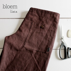 Puede incluir: Una prenda de lino marr&oacute;n con un bolsillo grande en la parte delantera. La prenda tiene un bot&oacute;n y una peque&ntilde;a etiqueta que dice "bloem linen".
