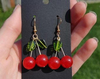 May include: A pair of drop earrings featuring two red spherical beads resembling cherries, attached to gold-toned metal hooks. Each earring includes a small green translucent leaf-shaped bead accent, suspended from the gold wire stems.