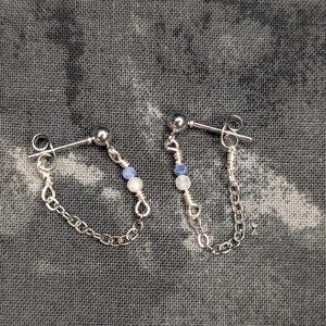 Puede incluir: Un par de pendientes de botón plateados con una delicada cadena que conecta el poste con la base. Cada pendiente está decorado con una pequeña cuenta azul y una pequeña cuenta blanca, aseguradas con alambre en la cadena de metal plateado.