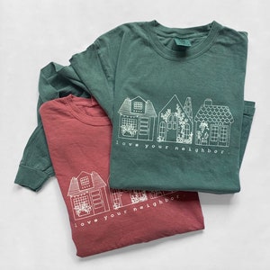 May include: Two long-sleeved t-shirts in shades of green and red. Each shirt features a white line drawing of houses with the text "love your neighbor." The shirts are made of a soft material and are suitable for casual wear.
