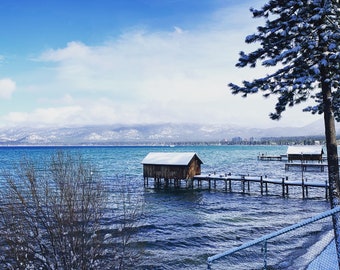 May include: A snowy winter scene with a wooden cabin on a pier over a blue lake. The cabin has a snow-covered roof and is surrounded by snow-covered trees. The mountains in the background are also covered in snow.
