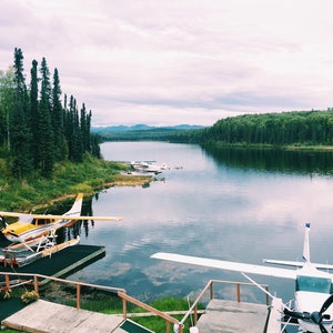 May include: A yellow and white seaplane docked at a wooden pier on a lake. The lake is surrounded by green trees and mountains in the distance.